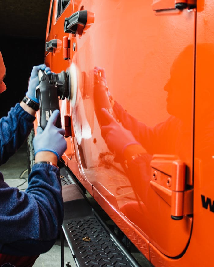 An adult male worker polishes an orange car with a power tool, reflecting dedication to detail.